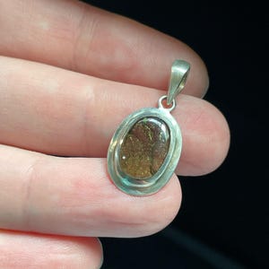 Puede incluir: Un colgante de plata con una piedra ovalada marr&oacute;n engastada en un bisel de plata. La piedra tiene una calidad brillante e iridiscente.