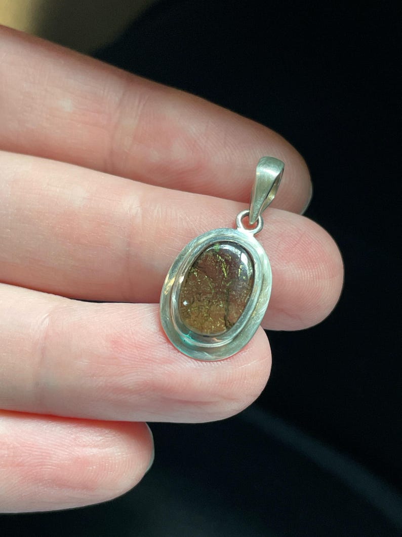 Puede incluir: Un colgante de plata con una piedra ovalada marr&oacute;n engastada en un bisel de plata. La piedra tiene una calidad brillante e iridiscente.