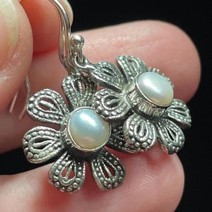 May include: Silver flower earrings with a pearl center. The earrings have a detailed, openwork design and a hook closure. The pearls are a soft, creamy white color. The earrings are shown against a dark background.