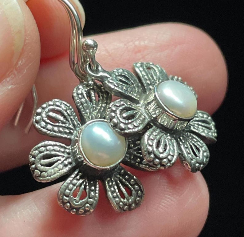 May include: Silver flower earrings with a pearl center. The earrings have a detailed, openwork design and a hook closure. The pearls are a soft, creamy white color. The earrings are shown against a dark background.