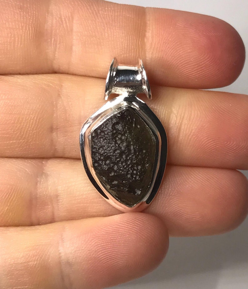 May include: A teardrop-shaped pendant featuring a dark brown, textured moldavite stone set in a polished silver frame. The pendant includes a silver bail at the top for attachment to a chain, showcasing a contrast between the rough stone and metal.