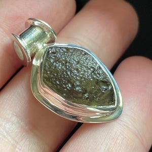 May include: A pendant featuring a rough, dark olive-green textured stone set in a polished silver bezel. The stone has an irregular, pitted surface and is encased in a smooth, teardrop-shaped silver frame with a cylindrical bail at the top.