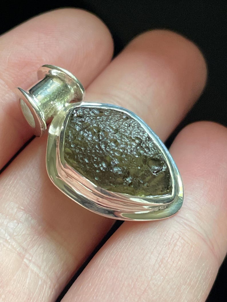 May include: A pendant featuring a rough, dark olive-green textured stone set in a polished silver bezel. The stone has an irregular, pitted surface and is encased in a smooth, teardrop-shaped silver frame with a cylindrical bail at the top.