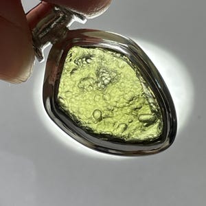 May include: A pendant featuring an irregular, translucent olive-green stone with a textured, bubbly surface set within a polished silver-toned metal frame. The stone is backlit, highlighting its natural inclusions and vibrant green color.