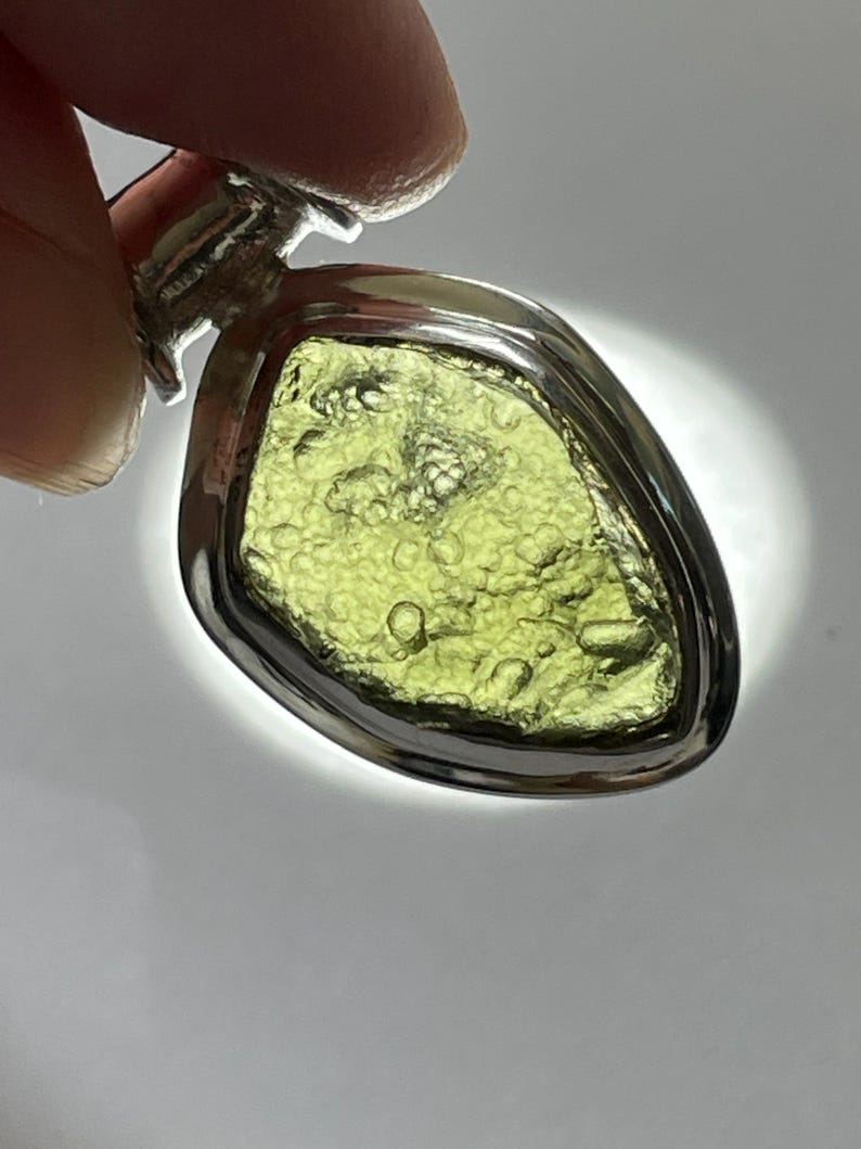 May include: A pendant featuring an irregular, translucent olive-green stone with a textured, bubbly surface set within a polished silver-toned metal frame. The stone is backlit, highlighting its natural inclusions and vibrant green color.