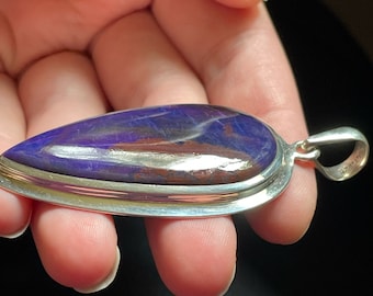 May include: A teardrop-shaped pendant with a purple and copper gemstone set in a silver bezel. The pendant has a silver bail.
