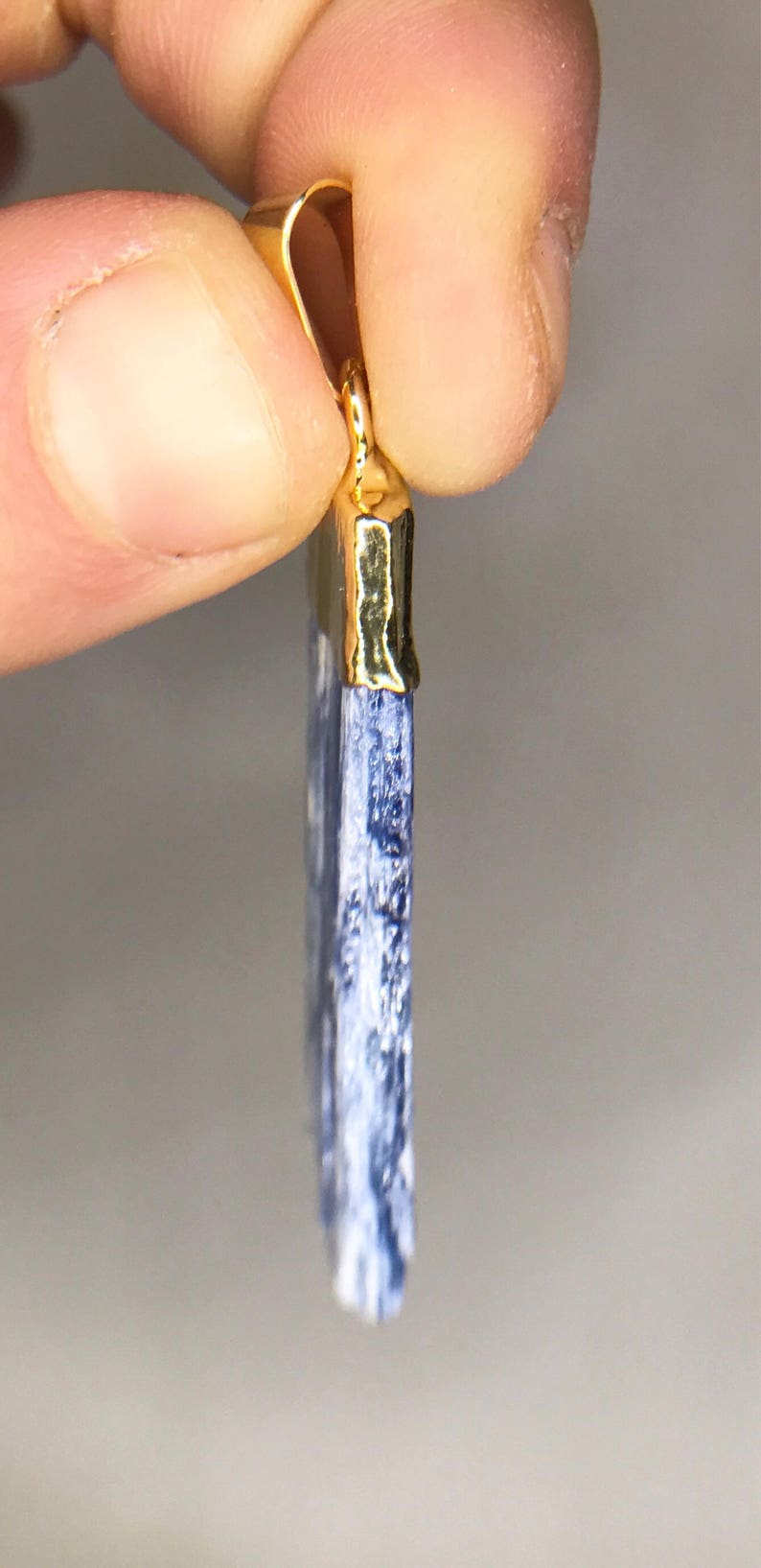 May include: A blue kyanite pendant with a gold-plated bail. The pendant is a long, thin rectangle with a pointed end.