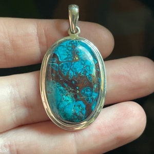 Peut inclure: Un pendentif en argent avec une grande pierre turquoise de forme ovale. La pierre pr&eacute;sente un motif tourbillonnant de bleu, de vert et de brun.
