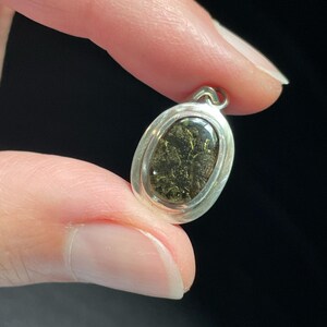 Puede incluir: Un colgante ovalado de plata con una piedra verde oscura engastada en el centro. La piedra tiene una textura brillante y reluciente.