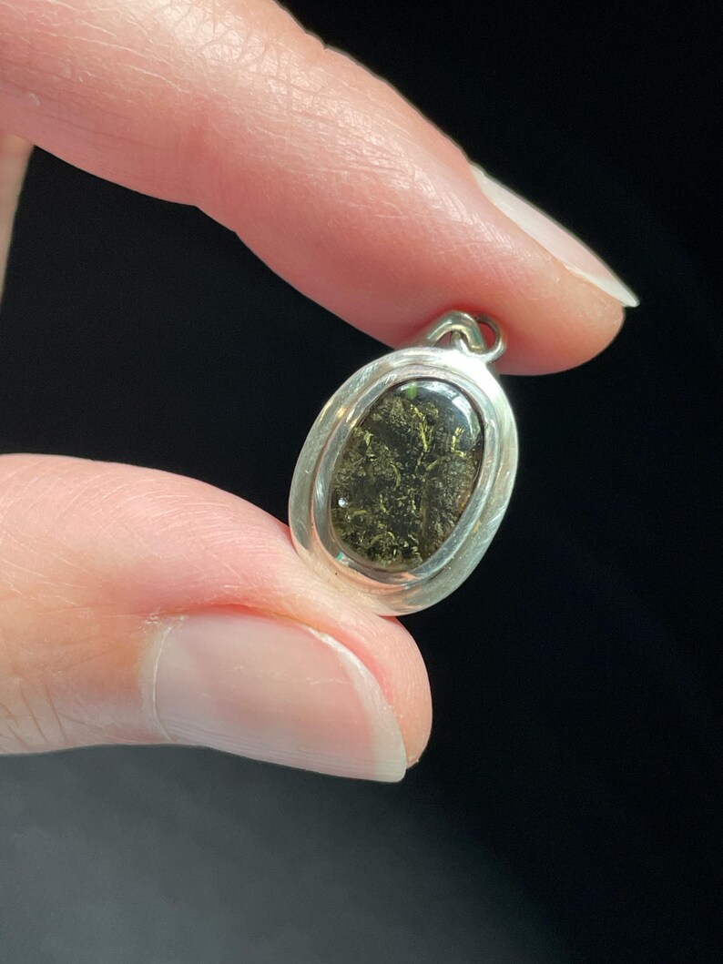Puede incluir: Un colgante ovalado de plata con una piedra verde oscura engastada en el centro. La piedra tiene una textura brillante y reluciente.