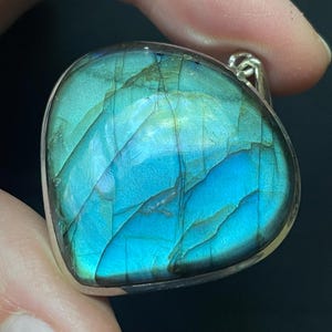May include: A heart-shaped labradorite pendant set in a silver frame. The gemstone displays a vibrant blue and teal iridescent flash across its polished surface, featuring natural internal fractures and a smooth, rounded contour with a metal bail.