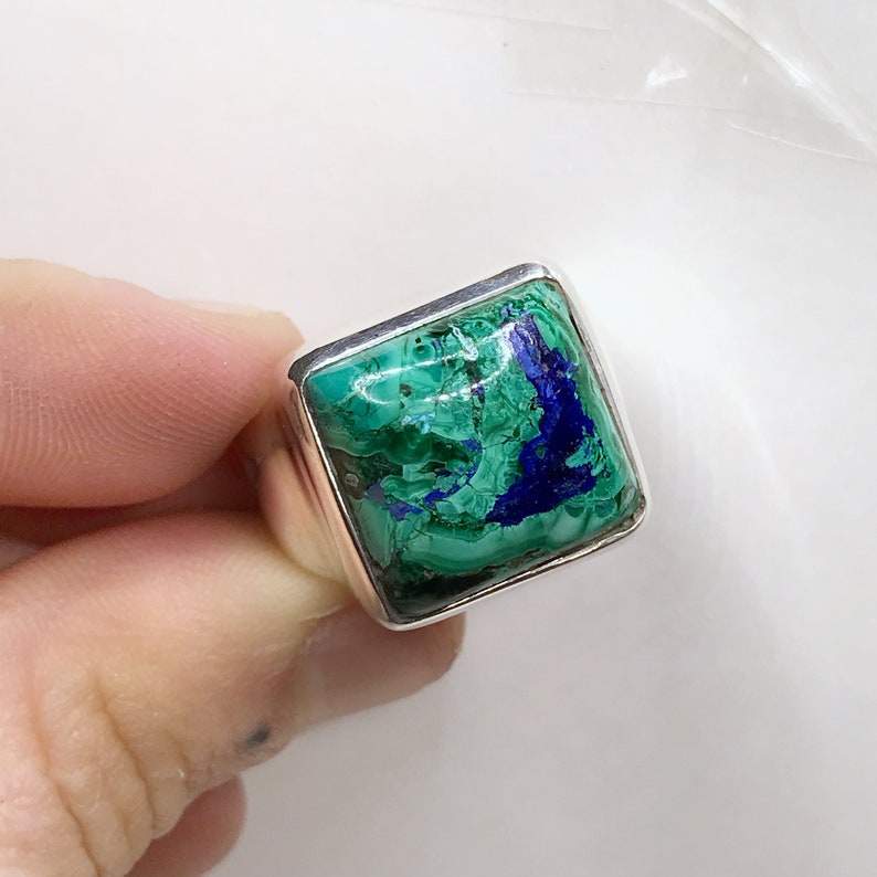 K&ouml;nnte beinhalten: Ein quadratischer Ring mit einer polierten Silberschiene und einem gro&szlig;en, flachen Edelstein mit wirbelnden Mustern aus leuchtend gr&uuml;nem Malachit und tiefblauem Azurit. Der Stein ist in einer glatten, schlichten Silberfassung eingefasst.