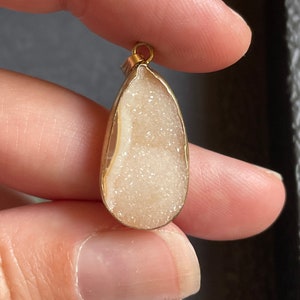 Puede incluir: Un colgante en forma de l&aacute;grima con un marco de metal dorado y una piedra blanca brillante de tipo cristal en el interior.