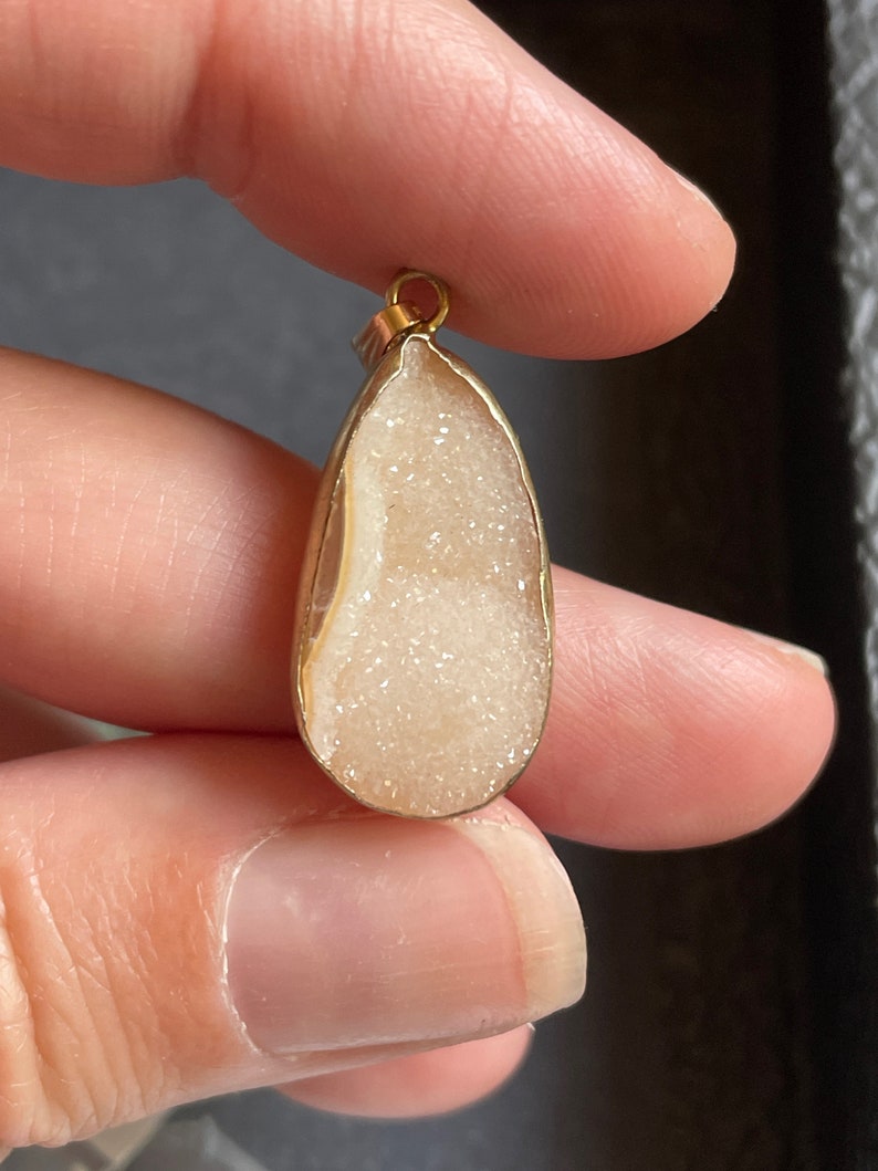 Puede incluir: Un colgante en forma de l&aacute;grima con un marco de metal dorado y una piedra blanca brillante de tipo cristal en el interior.