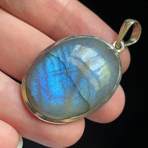 May include: An oval-shaped labradorite pendant set in a polished silver frame. The gemstone displays a prominent blue iridescent flash across its surface, featuring natural internal fractures and a smooth, rounded cabochon cut with a metal bail.