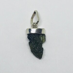 May include: A dark green, rough, and irregularly shaped stone pendant set in a silver bezel. The pendant is attached to a silver bail.