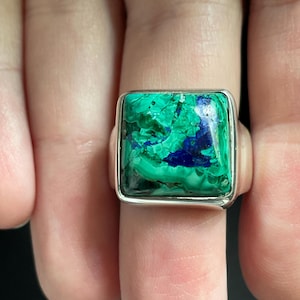 May include: A square-shaped ring featuring a polished silver band and a large, flat-topped gemstone with swirling patterns of vibrant green malachite and deep blue azurite, set within a smooth, simple silver bezel frame.