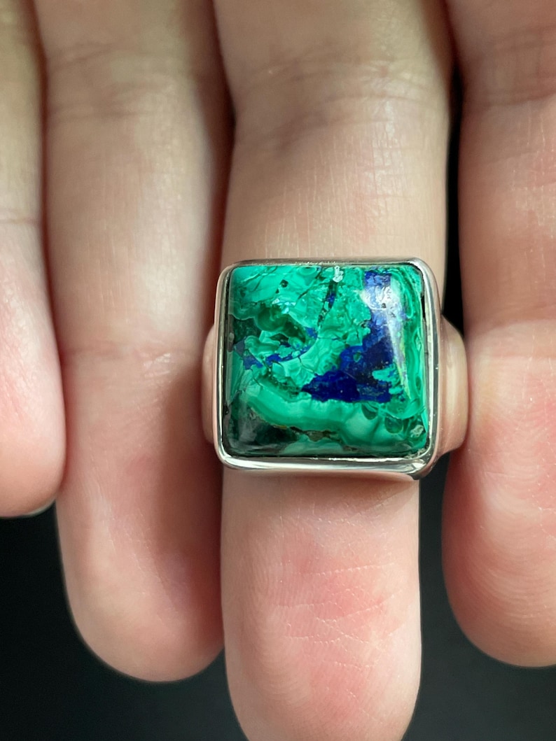 K&ouml;nnte beinhalten: Ein quadratischer Ring mit einer polierten Silberschiene und einem gro&szlig;en, flachen Edelstein, der wirbelnde Muster aus leuchtend gr&uuml;nem Malachit und tiefblauem Azurit aufweist und in einer glatten, einfachen Silberfassung sitzt.