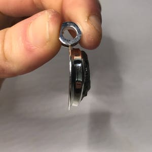 May include: A side profile view of a pendant featuring a dark, textured, irregular stone set within a polished, reflective silver-toned metal frame. The pendant includes a circular bail at the top for attachment to a chain or cord, showcasing the contrast between the smooth metallic edge and the rough, dark surface of the stone.