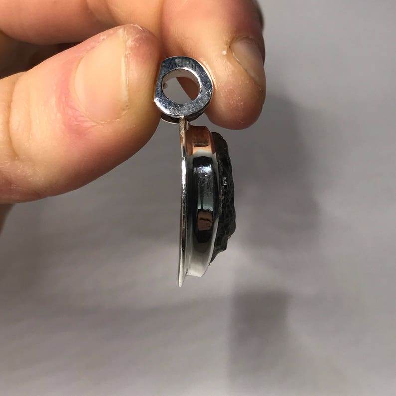 May include: A side profile view of a pendant featuring a dark, textured, irregular stone set within a polished, reflective silver-toned metal frame. The pendant includes a circular bail at the top for attachment to a chain or cord, showcasing the contrast between the smooth metallic edge and the rough, dark surface of the stone.