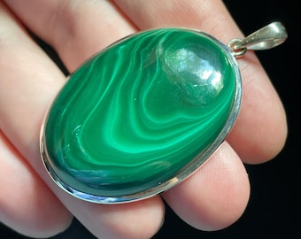 May include: A green malachite oval pendant with a silver setting. The pendant is held in a hand.