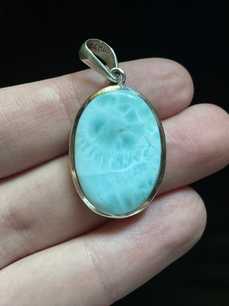 Puede incluir: Un colgante de plata con una piedra ovalada de color azul claro. La piedra tiene un patr&oacute;n de remolino y nublado.