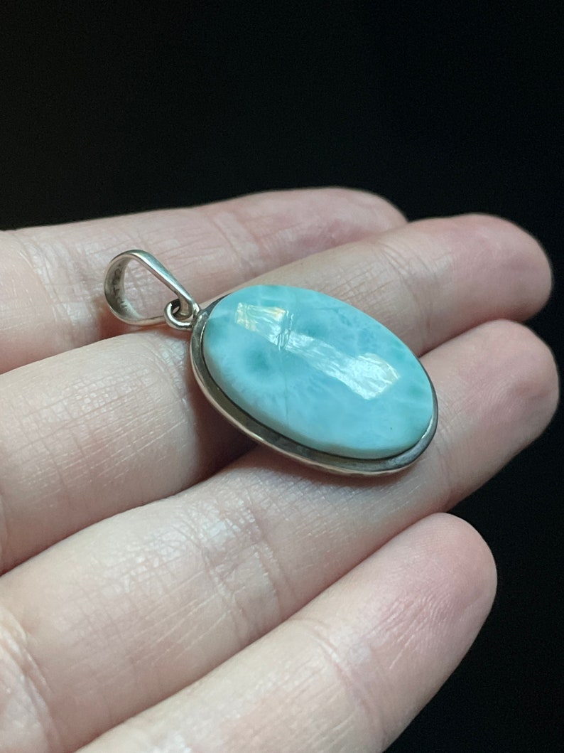 Puede incluir: Un colgante de plata con una piedra ovalada de color azul claro. La piedra tiene un aspecto nublado y veteado.