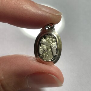 Puede incluir: Un colgante ovalado de plata con un dise&ntilde;o texturizado de color plateado en el interior. El colgante tiene un bucle en la parte superior para sujetarlo a una cadena.