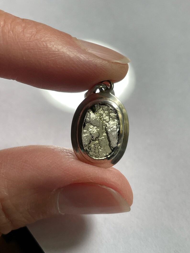 Puede incluir: Un colgante ovalado de plata con un dise&ntilde;o texturizado de color plateado en el interior. El colgante tiene un bucle en la parte superior para sujetarlo a una cadena.