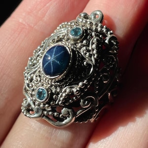 May include: A silver ring with a blue star sapphire and two smaller blue gemstones. The ring has a detailed, ornate design.