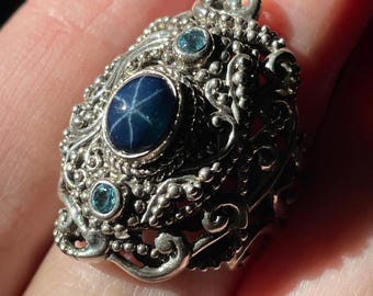 Könnte beinhalten: Ein silberner Ring mit einem blauen Stern-Saphir und zwei kleineren blauen Edelsteinen. Der Ring hat ein detailliertes, verziertes Design.