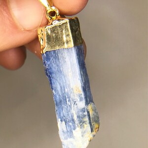 May include: A blue kyanite crystal pendant with a gold-toned setting. The crystal is a long, thin, and slightly irregular shape.