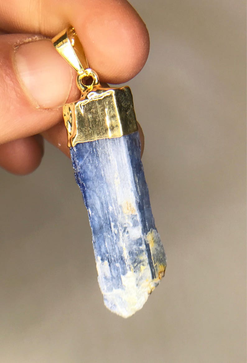 May include: A blue kyanite crystal pendant with a gold-toned setting. The crystal is a long, thin, and slightly irregular shape.