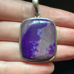 May include: A rectangular pendant with a vibrant purple stone, set in a silver-colored metal frame. The stone has a unique pattern with shades of purple and gray. The pendant has a silver loop for a chain.