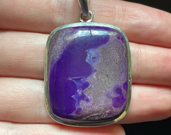 May include: A rectangular pendant with a vibrant purple stone, set in a silver-colored metal frame. The stone has a unique pattern with shades of purple and gray. The pendant has a silver loop for a chain.
