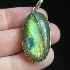 May include: An oval-shaped labradorite gemstone pendant set in a polished silver-toned metal bezel. The stone displays iridescent flashes of lime green, yellow, and deep blue across its smooth, reflective surface, topped with a silver-toned bail.