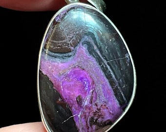 May include: A silver pendant featuring a purple and black stone with swirling patterns. The stone is set in a silver bail.