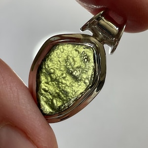 May include: A pendant featuring a rough, textured, olive-green moldavite stone set within a polished silver bezel. The stone has an irregular, pitted surface with small bumps and depressions, held by a smooth, shiny silver frame with a bail at the top.