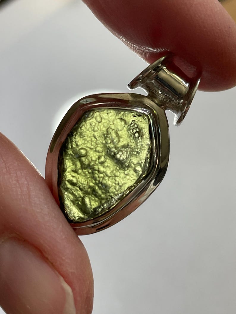 May include: A pendant featuring a rough, textured, olive-green moldavite stone set within a polished silver bezel. The stone has an irregular, pitted surface with small bumps and depressions, held by a smooth, shiny silver frame with a bail at the top.