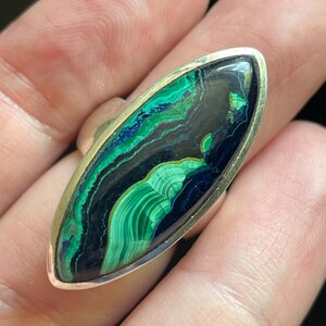 May include: A marquise-shaped ring featuring a polished malachite and azurite gemstone with swirling bands of deep green, bright green, and dark blue. The stone is set in a smooth, plain silver metal bezel that follows the elongated, pointed shape.