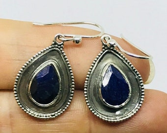 May include: A pair of silver-toned teardrop earrings. Each earring features a dark blue, pear-shaped gemstone set within a textured frame. The earrings have a beaded border and a simple hook closure.