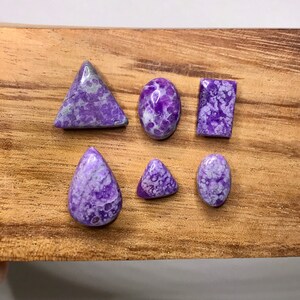 May include: Six polished gemstones in various shapes: a triangle, oval, rectangle, teardrop, small triangle, and oval. The stones are a vibrant purple with white and gray marbling, set against a wooden surface.