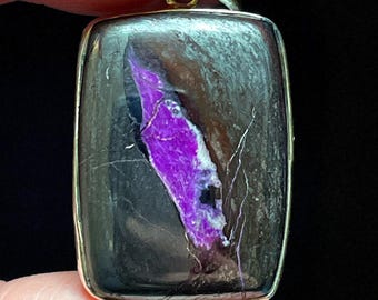 May include: Rectangular pendant with a silver-colored frame. The stone features a striking contrast of deep purple and black hues, with delicate white veining. The pendant is held by a finger.