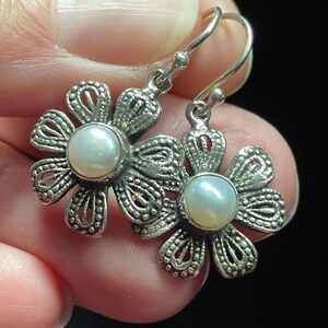 May include: A pair of silver-toned flower earrings with a pearl center. The earrings have a detailed, openwork design and a hook closure. The earrings are held in a hand against a dark background.