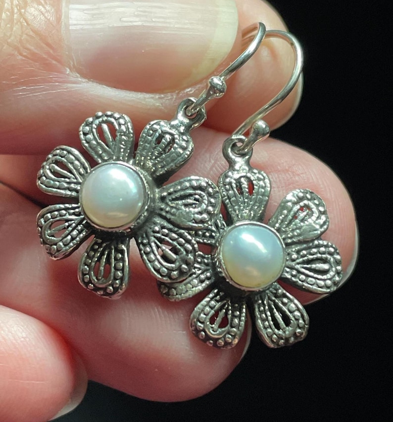 May include: A pair of silver-toned flower earrings with a pearl center. The earrings have a detailed, openwork design and a hook closure. The earrings are held in a hand against a dark background.