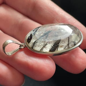 May include: A silver pendant featuring a clear quartz crystal with black inclusions. The pendant is oval shaped and has a smooth, polished finish.