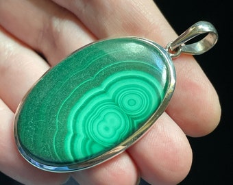 May include: A silver pendant featuring a green malachite stone with a swirling pattern. The stone is oval shaped and set in a silver bezel.