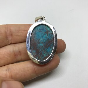 Peut inclure: Un pendentif ovale en argent avec une pierre turquoise sertie au centre. La pierre pr&eacute;sente un motif marbr&eacute; bleu et brun.