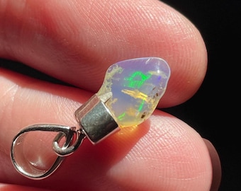 May include: A raw, translucent opal pendant set in a polished silver-toned metal cap with a circular bail. The stone displays iridescent flashes of green, yellow, and blue light against a milky, pale base, showcasing its natural, irregular shape.