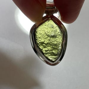 May include: A pendant featuring a raw, textured, olive-green moldavite stone set in a polished silver-toned metal frame. The stone has a pitted, irregular surface with a translucent quality, held within a smooth, teardrop-shaped metal bezel setting.